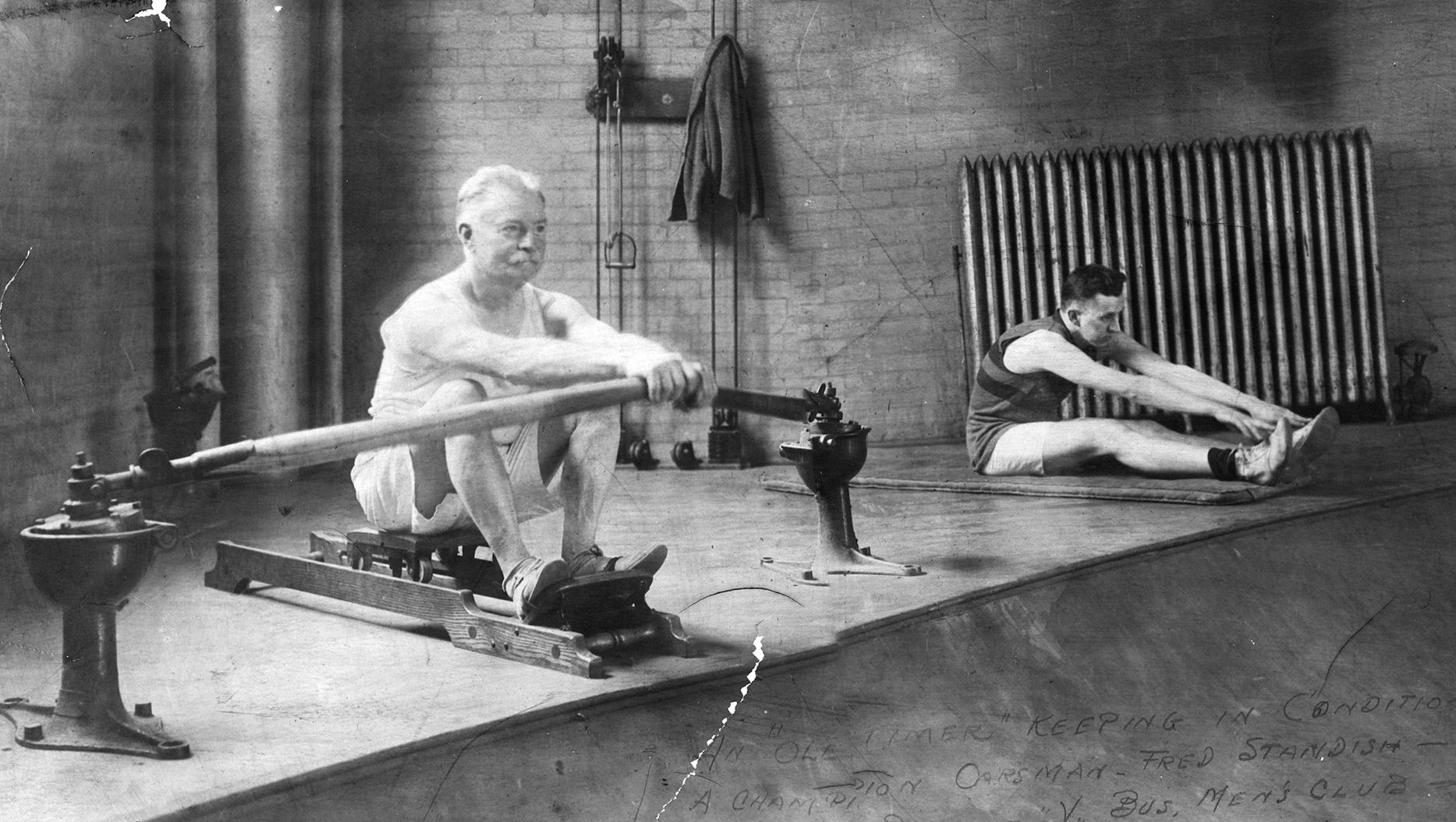 An older man uses a rowing machine at Detroit's YMCA, while a younger man stretches. Handwritten on the photograph: "An 'ole timer' keeping in condition. A champion oarsman, Fred Standish, member Detroit Y. Both men were members of the Business Men's Club at the YMCA started by A.G. Studer."