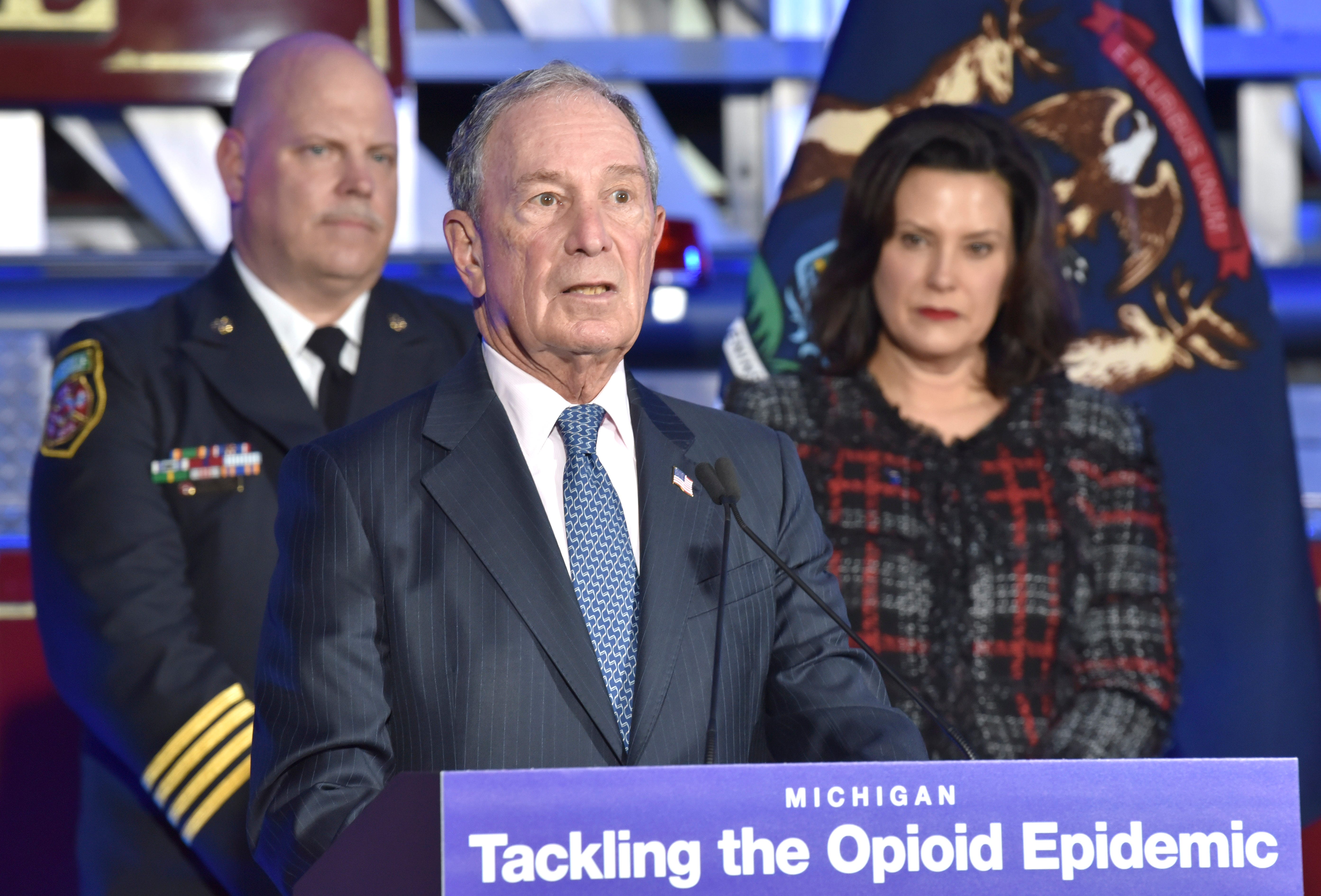 Former City of New York Mayor Michael Bloomberg speaks from the podium as Eastpointe Deputy Fire Chief Nick Sage, left, and Michigan Governor Gretchen Whitmer, right, listen.
