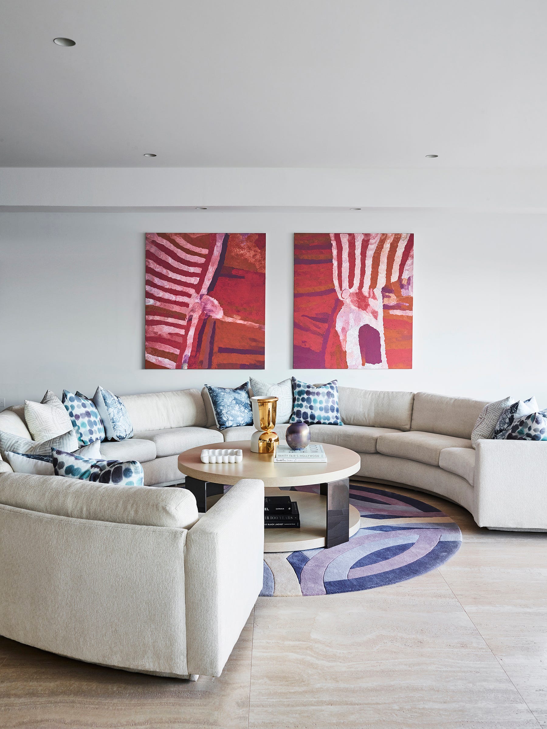 A sculpted circular rug punctuated by shades of lavender follows the form of a curved sofa, accented with pillows in shades of blue. Adding a jolt in color and pattern is a pair of paintings by the indigenous Australian artist Biddee Baadjo.