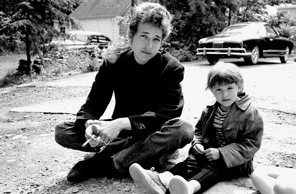 Bob Dylan sits in the driveway of the Cafe Espresso, Woodstock, NY 1964.  The young girl is the daughter of Bernard and May Lou Paturel, who own the Cafe Espresso.