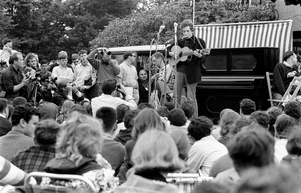 Bob Dylan performing at Newport Folk Festival, Newport, Rhode Island, 1964. Pete Seeger is in the chair to right of piano.