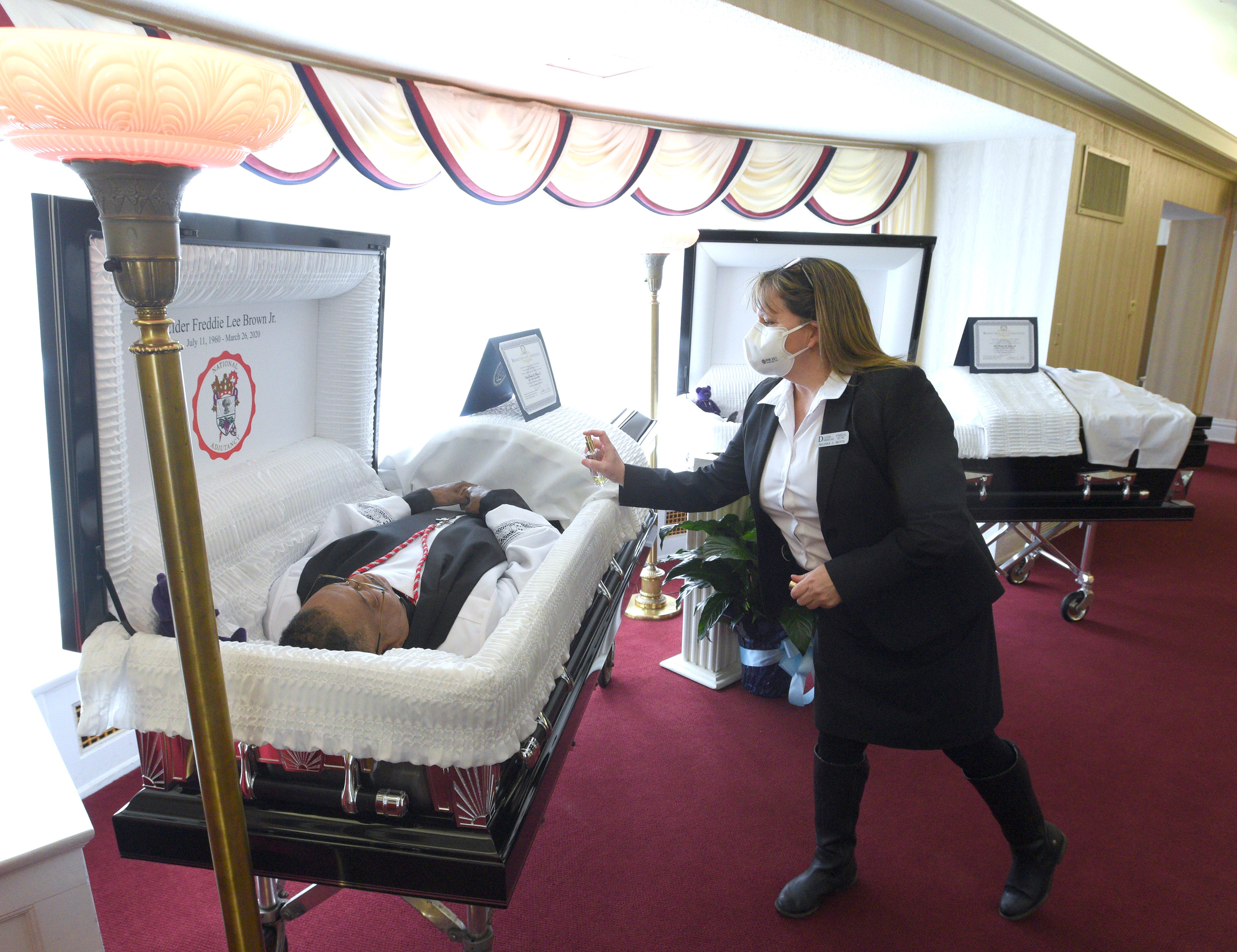 Following the wishes of wife and mother Sandy Brown (not pictured), funeral home owner Melissa Butts sprays Freddie Lee Brown Jr. ' s casket with the cologne Aramis, Brown Jr. ' s favorite. Butts also sprayed Freddie Lee Brown III ' s casket with his favorite fragrance, Guilty by Gucci. The father and son died three days apart from COVID-19 and are laid to rest together on Friday, April 10, 2020.