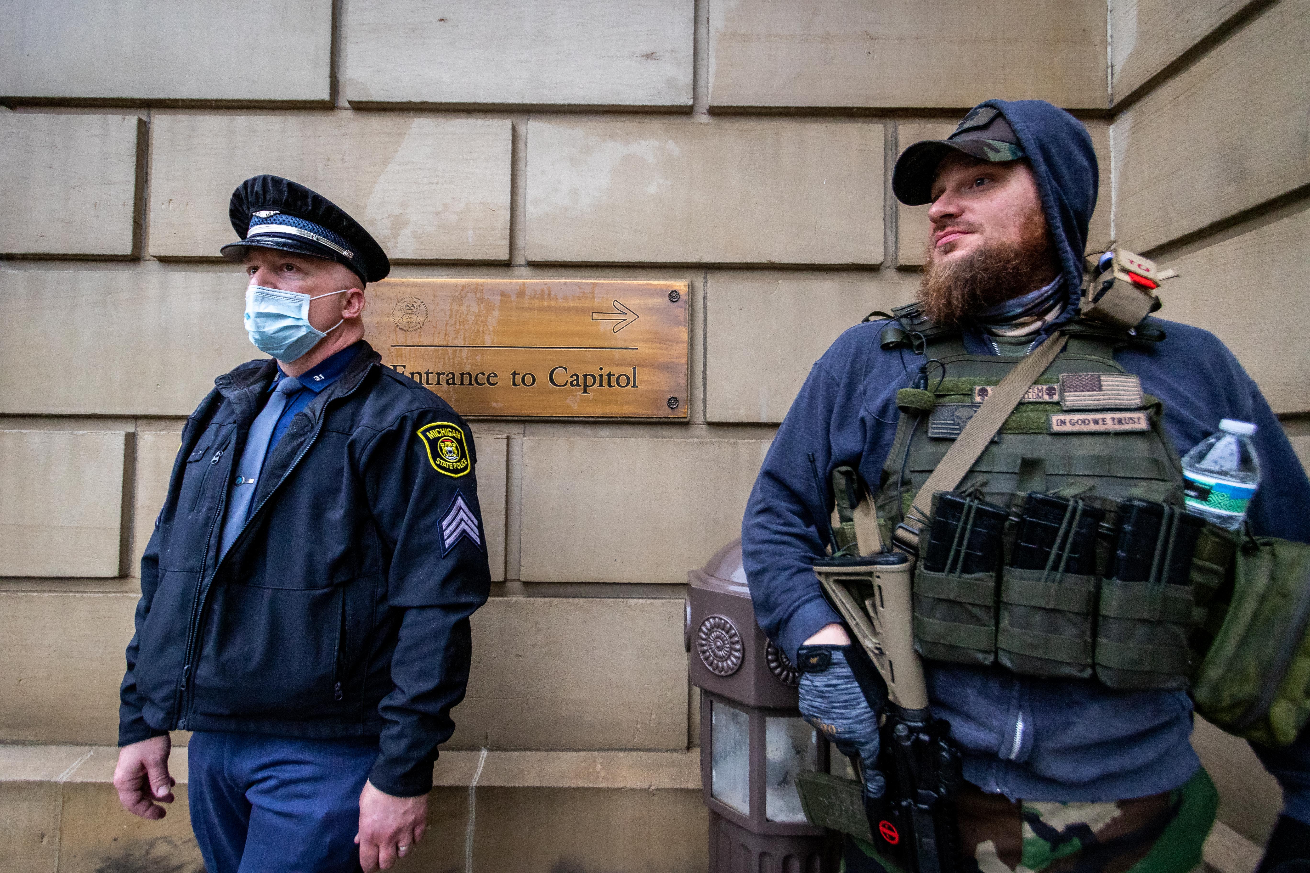 Michigan State trooper stands guard at the lower entrance to state Capitol building beside a protestors strapped with deadly weapons.